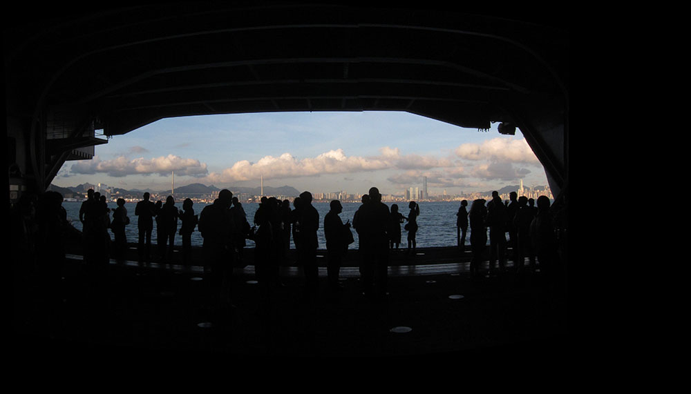 Photo of Elevator Opening on a Large Aircraft Across a Harbor to a Large City, with People in Silhouette.